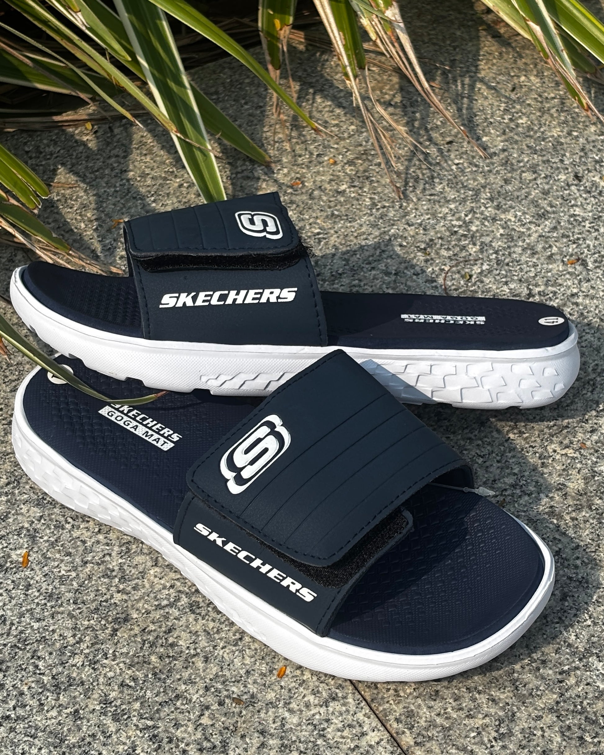 Navy and white Skechers sandals on a concrete surface with plants in the background