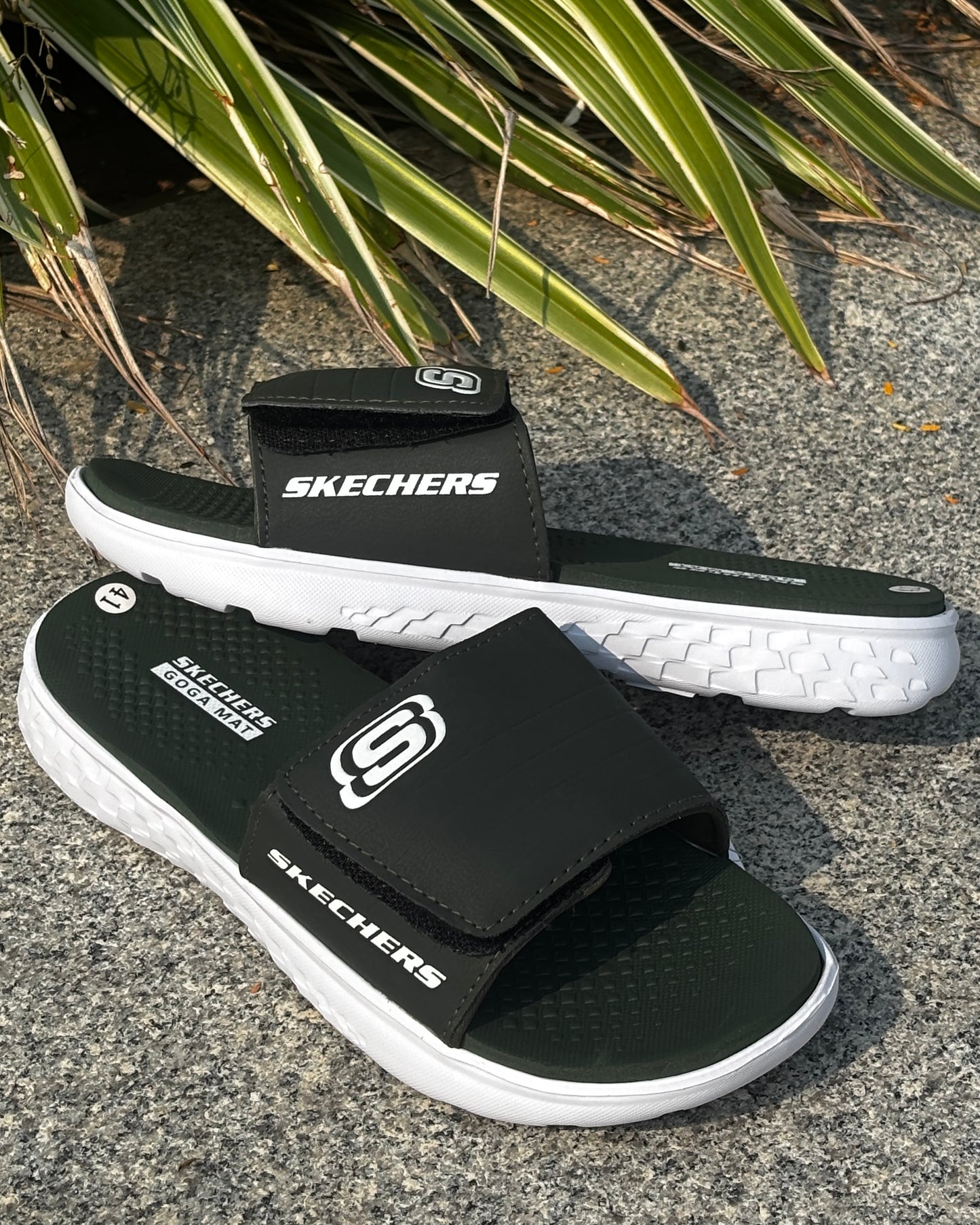 Olive and white Skechers sandals on a textured surface with green leaves in the background

